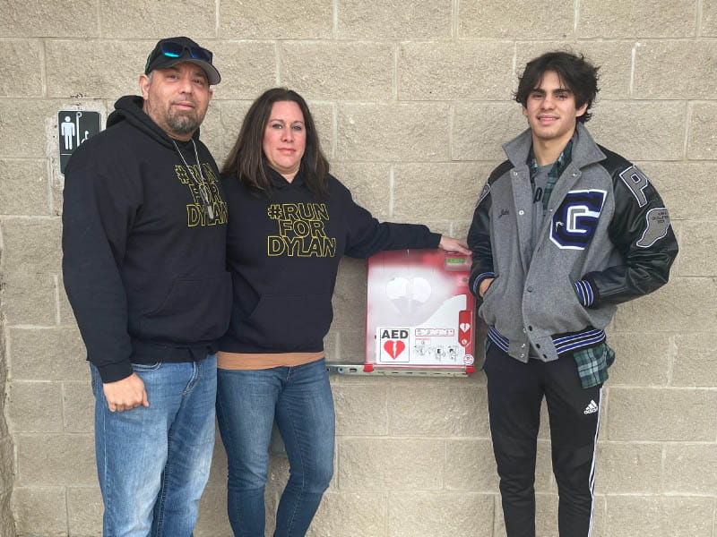 From left: Dylan's parents, Gus and Lisa Dorrell, and brother, Jake Dorrell, with the AED that was placed in the park where Dylan collapsed. (Photo courtesy of the Dorrell family)