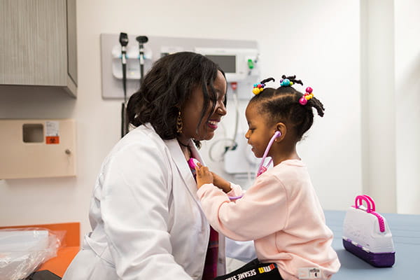 A young Black girl is using a stethoscope to listen to the heartbeat of her Black female doctor in a patient room.