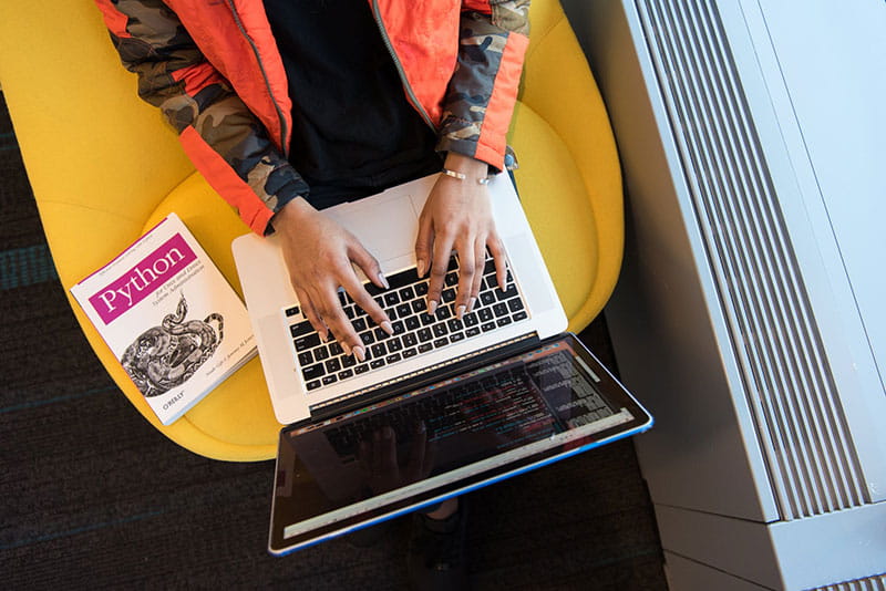 An aerial view of a person typing on a laptop with a Python coding language book on the seat next to them.